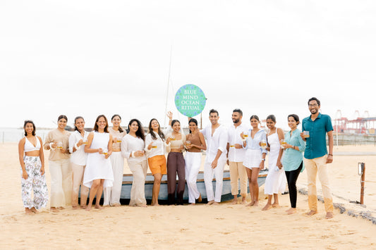 Group of people in light summer clothing holding drinks on a sandy beach near a 'Blue Mind Ocean Ritual' sign.